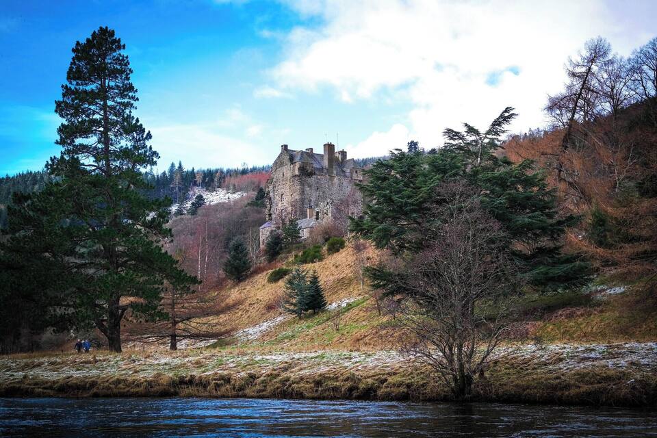 A historic stone castle on a hill, surrounded by trees and a river.
