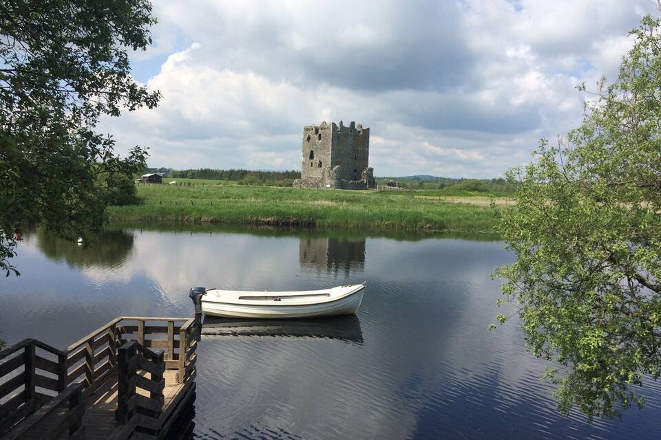 A calm river with a small boat docked at a wooden pier. In the background, there is a historic stone tower.