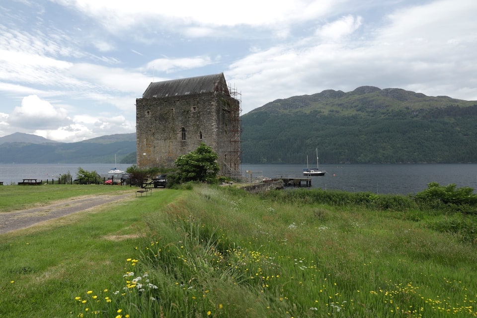 Ein historischer SteinTurm an einem See mit Booten, umgeben von üppiger Vegetation und Bergen.