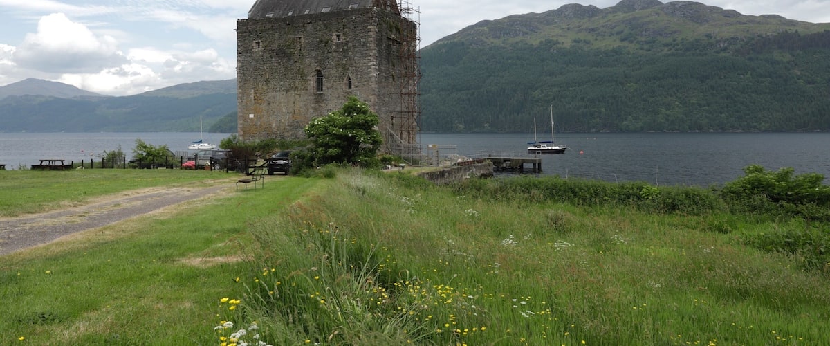 A historic stone tower by a lake with boats, surrounded by lush greenery and mountains.