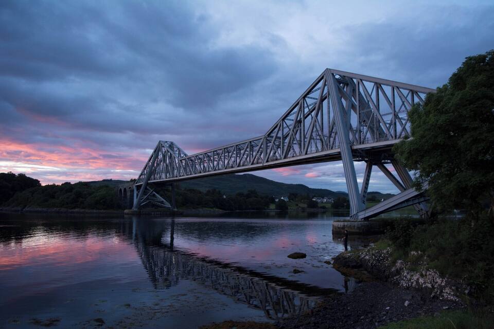 A steel bridge over a calm river at dusk with a pink sky.