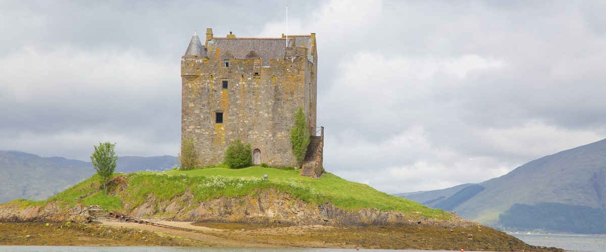 A historic stone tower on a grassy hill by a body of water, with mountains in the background.