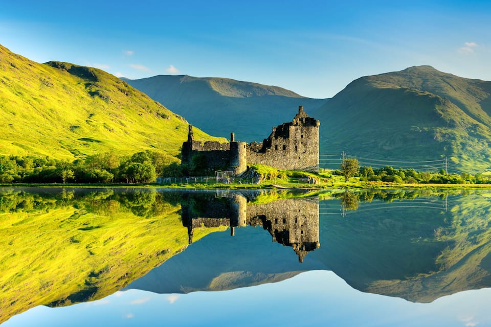 A castle on a hill overlooking a calm lake with mountains in the background.
