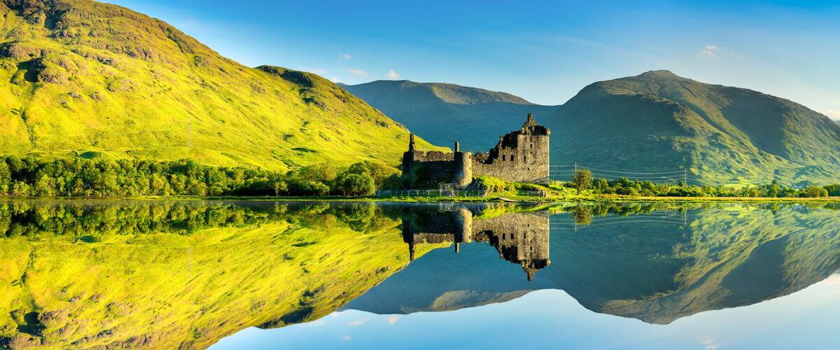 A castle on a hill overlooking a calm lake with mountains in the background.