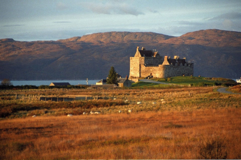 Um castelo histórico numa colina gramada, com um lago e montanhas ao fundo.