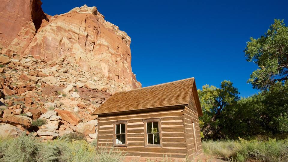 A wooden cabin with a red roof, surrounded by desert vegetation and a large red rock formation in the background.