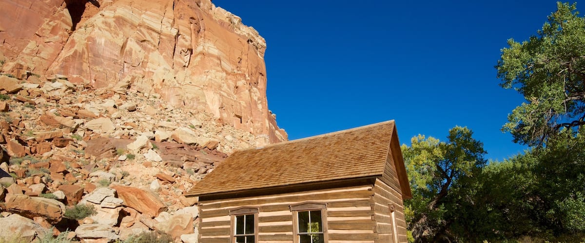 A wooden cabin with a red roof, surrounded by desert vegetation and a large red rock formation in the background.