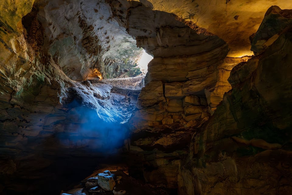 A cave with a bridge over a blue water pool, illuminated by artificial light.