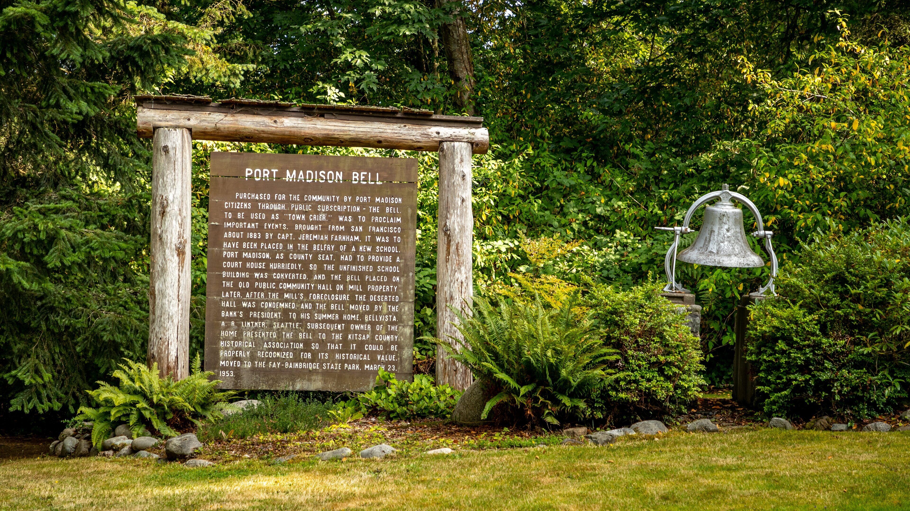 A wooden sign with historical information about the Port Madison Bell, surrounded by lush greenery and a large bell.