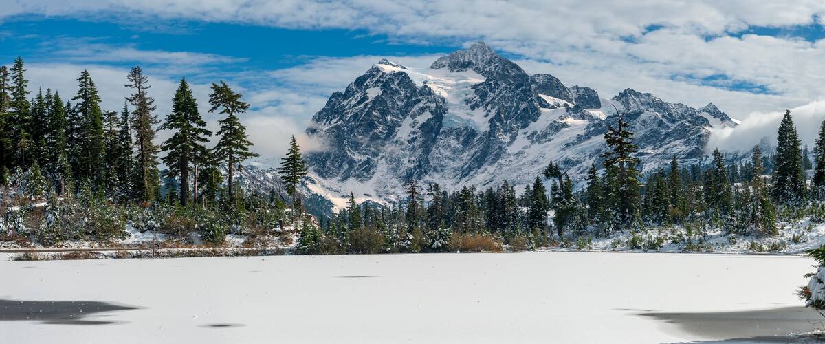 A snow-covered lake surrounded by dense evergreen trees and a snow-capped mountain range.