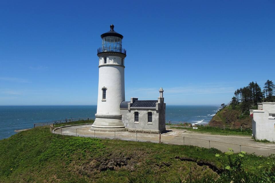 Ein weißer Leuchtturm mit schwarzer Spitze thront auf einem grasbewachsenen Hügel mit Blick auf das Meer.