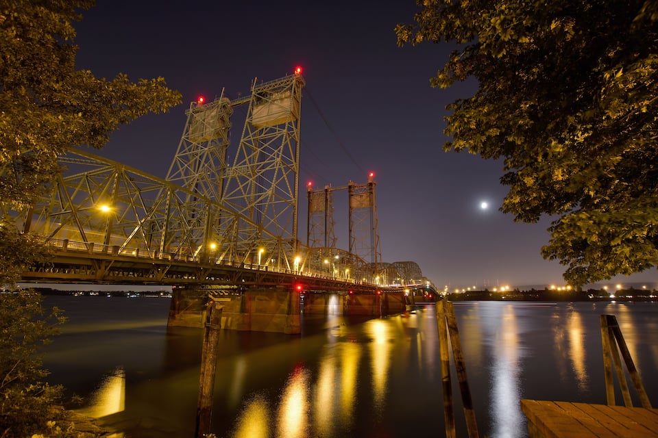 A nighttime view of a bridge with illuminated lights, a calm river below, and a tree on the right.