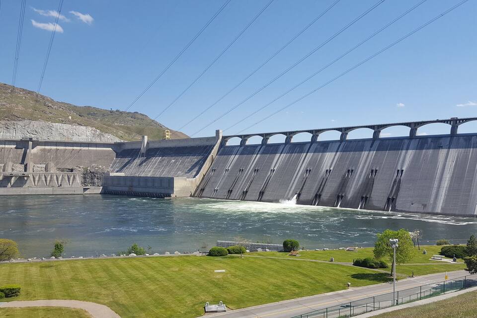 Een grote dam met stromend water, een grasrijk gebied op de voorgrond en een heuvellandschap op de achtergrond.