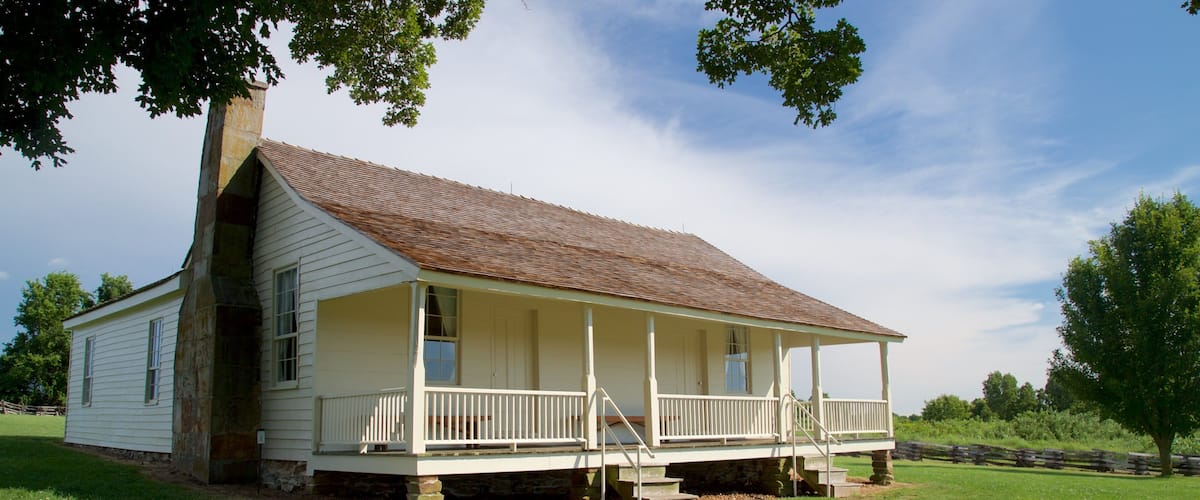 A white wooden house with a porch, surrounded by a grassy field and trees.