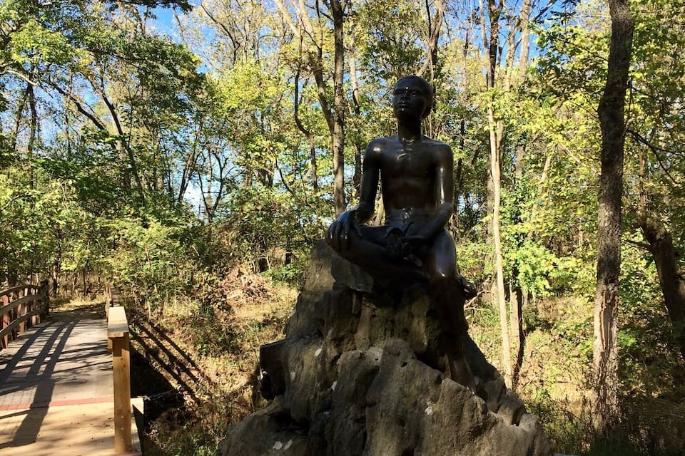 A statue of a seated figure on a rock, surrounded by trees and a wooden bridge.