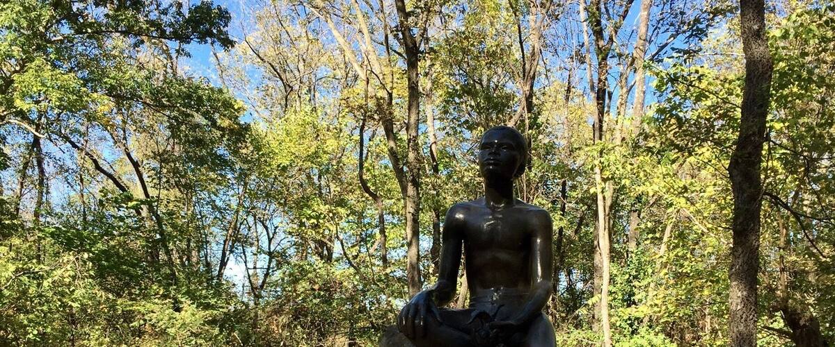 A statue of a seated figure on a rock, surrounded by trees and a wooden bridge.
