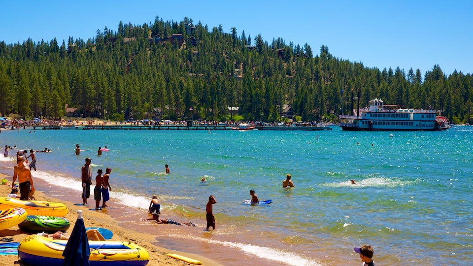 A beach with people swimming and playing, a boat, and a forested hill in the background.