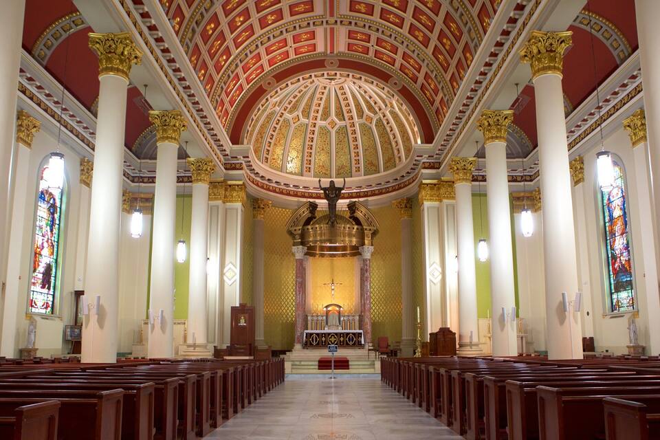A church interior with a high, ornate ceiling, stained glass windows, and rows of wooden pews.