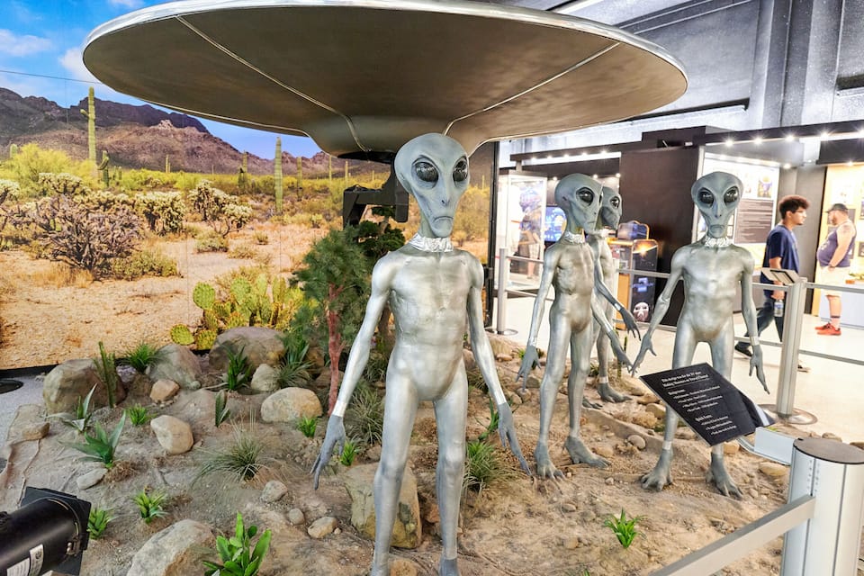 Three alien statues in a desert exhibit with a rocky landscape in the background.
