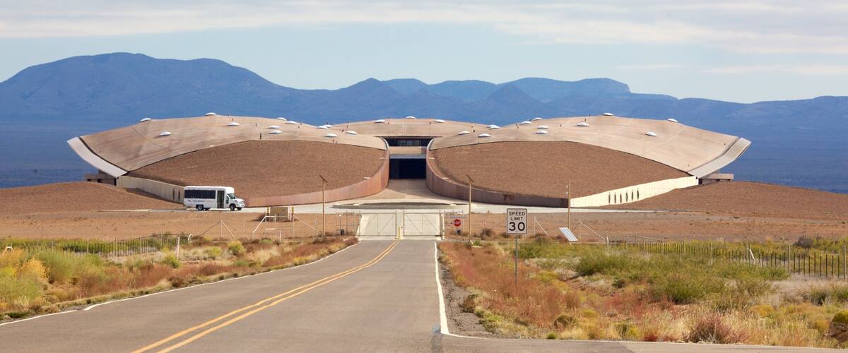 A large, unique building with a curved roof structure, a road leading to it, and mountains in the background.