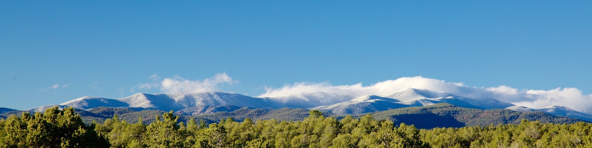 A mountain range with snow-capped peaks under a clear blue sky, surrounded by dense green forest.