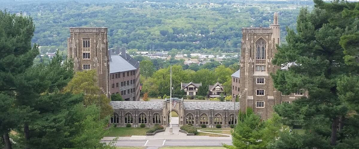 A large, historic building with two tall towers and a central archway, surrounded by lush green trees.