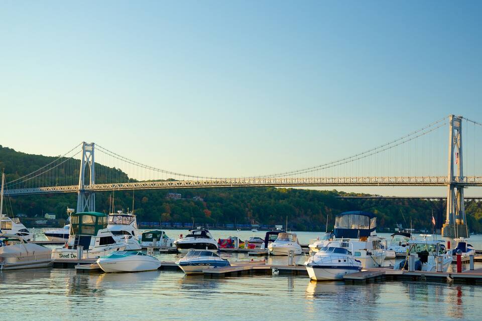 A marina with various boats docked, a large suspension bridge in the background, and a forested hillside.