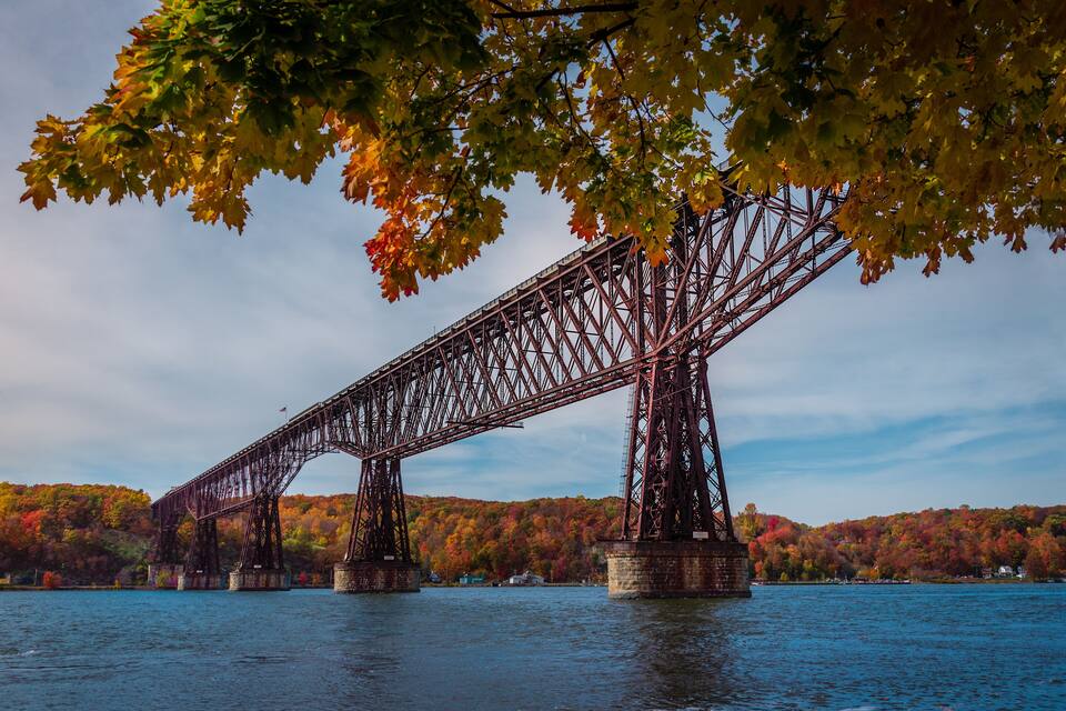 A steel bridge over a body of water with autumn foliage in the background.