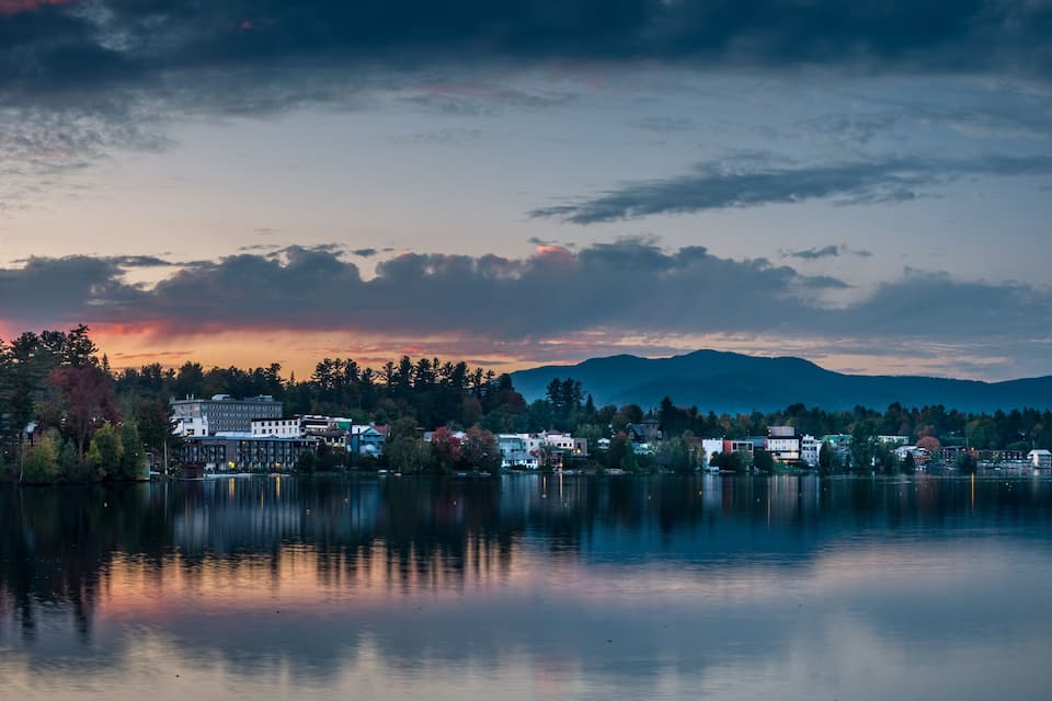 A calm lake reflecting a sunset with mountains in the background and a town with buildings along the shore.