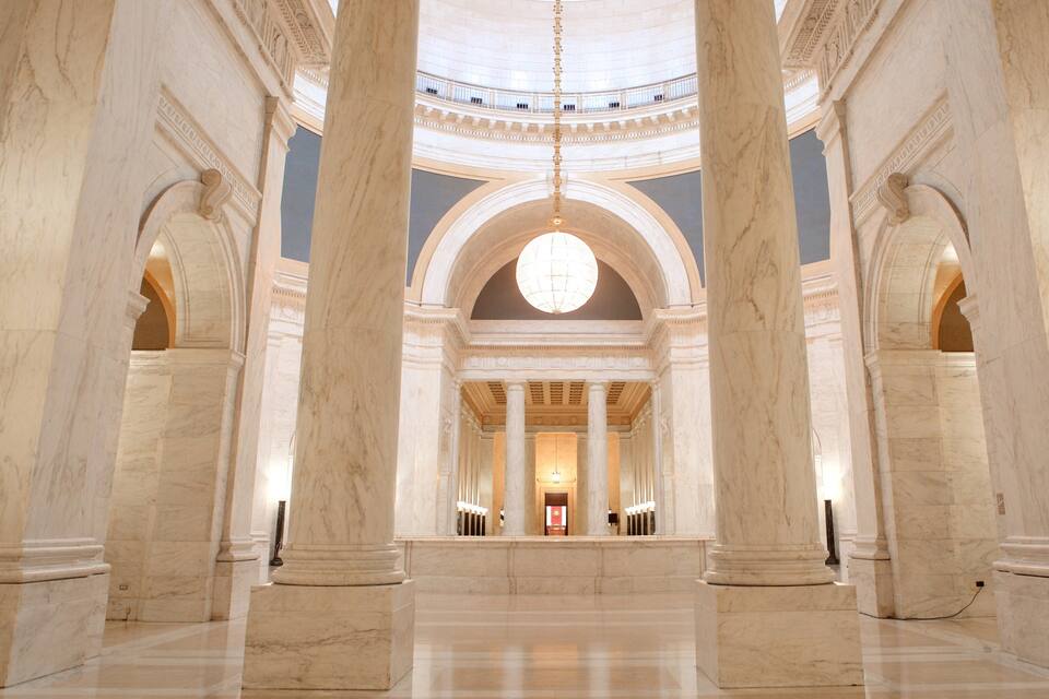 A grand hall with tall marble columns, a large circular skylight, and a reflective floor.