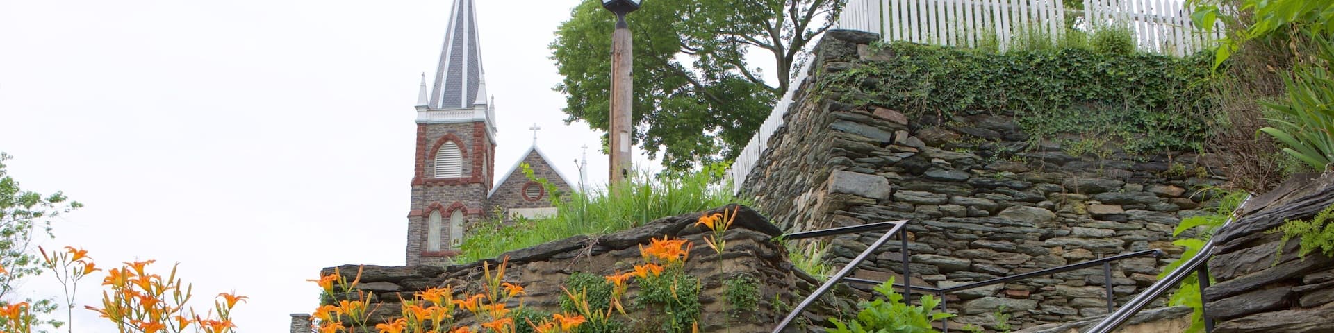Una escalera de piedra con flores anaranjadas, la aguja de una iglesia y una valla de piquetes blancos.