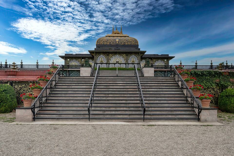 A grand staircase leading to a domed building with intricate golden patterns, surrounded by lush greenery and red flowers.