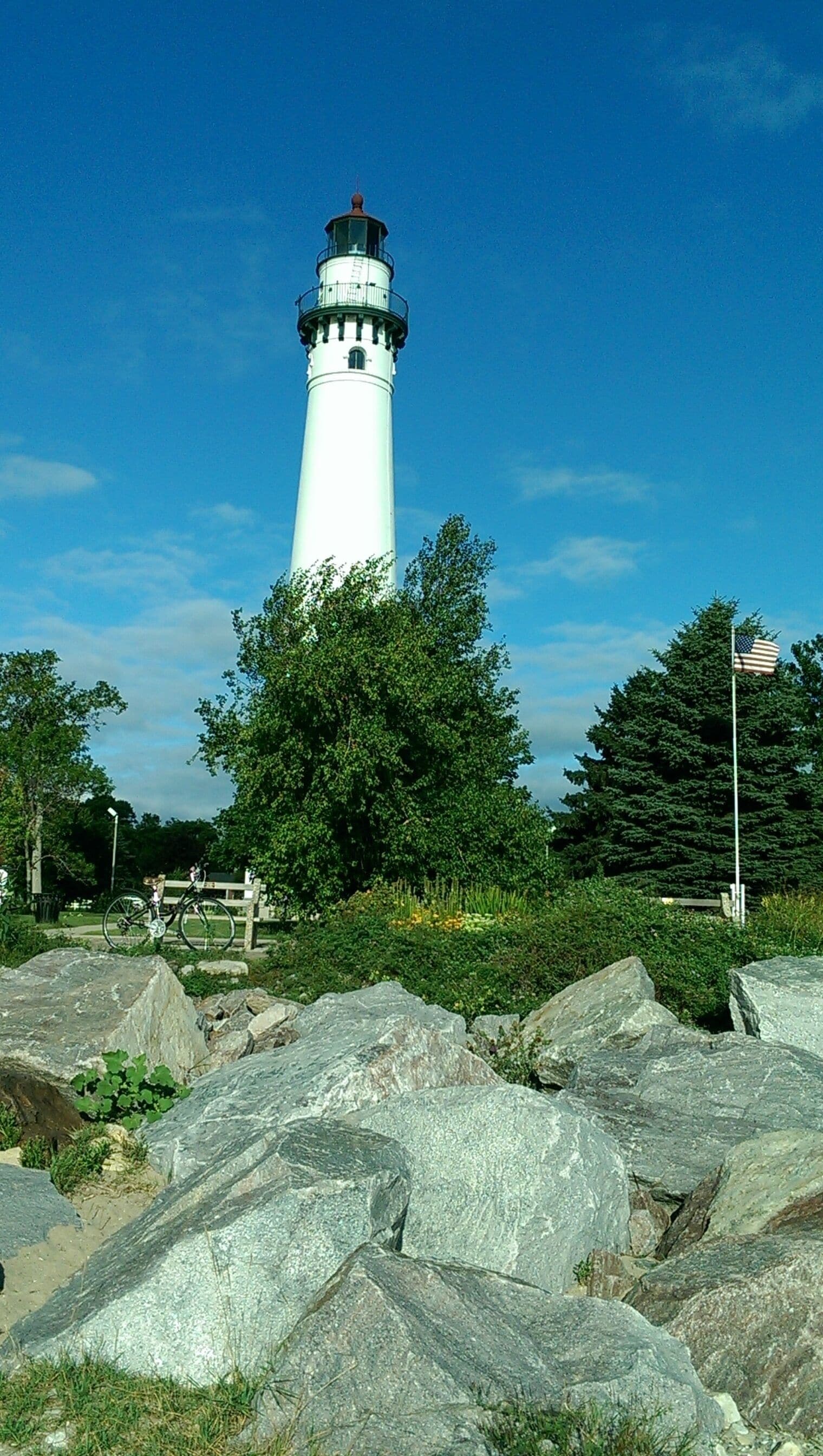 A white lighthouse with a black top, surrounded by large rocks and green trees.