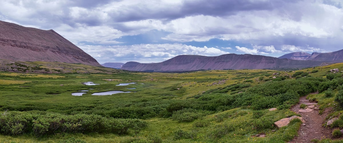 A green valley with small ponds, surrounded by mountains under a cloudy sky.