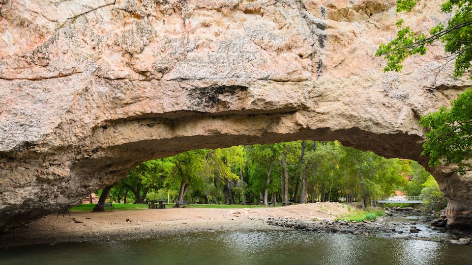 A large rock arch over a river with trees and a park in the background.