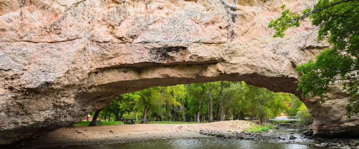 A large rock arch over a river with trees and a park in the background.