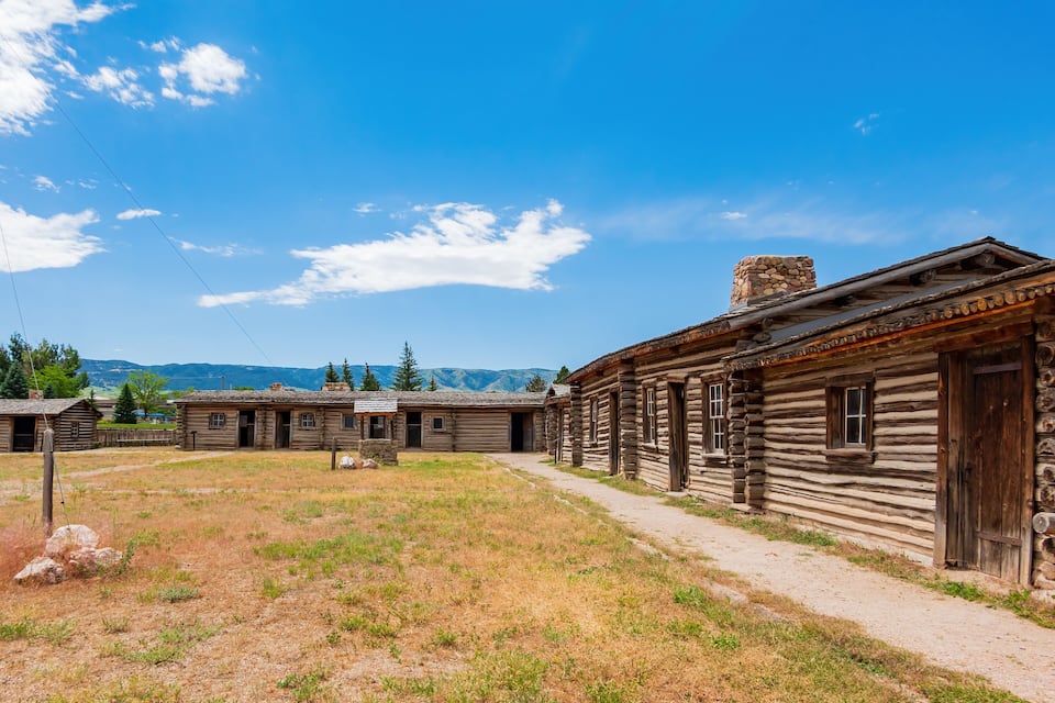 A rustic log cabin with a stone chimney, surrounded by a grassy field and a clear blue sky.
