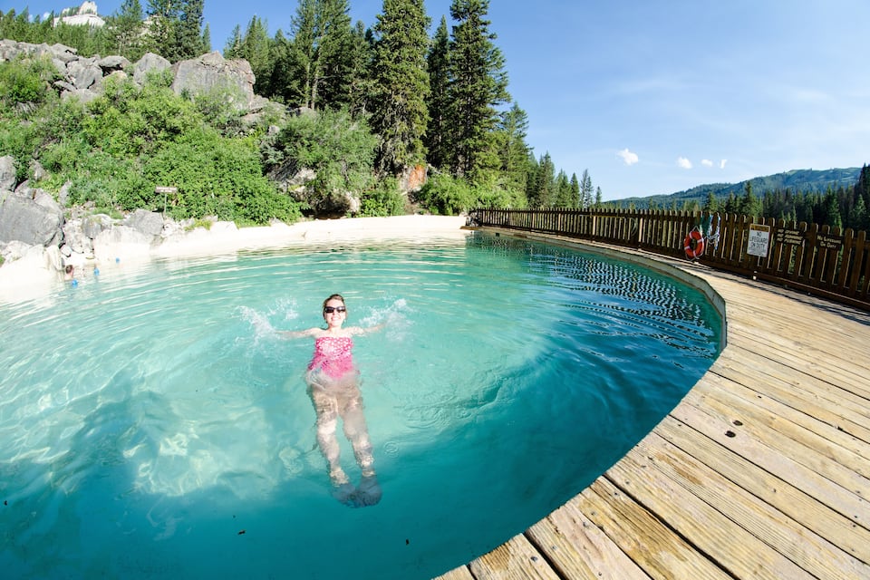 A person floating in a clear blue pool surrounded by a wooden deck, with a forested hillside and a wooden fence in the background.