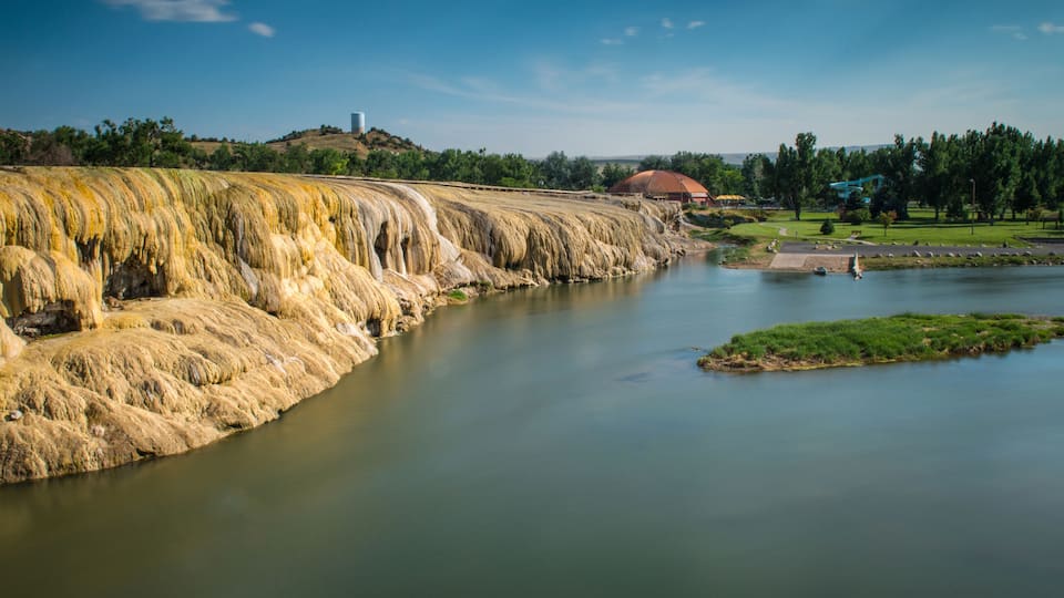 A river with a waterfall, a small island, and a building with a red roof in the background.