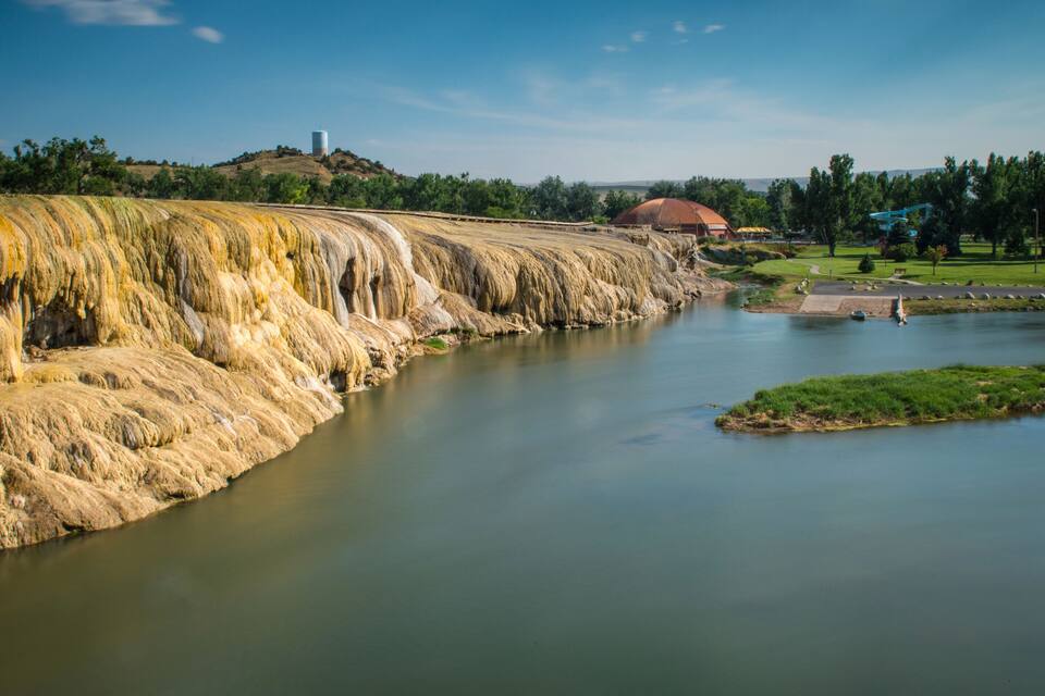 A river with a waterfall, a small island, and a building with a red roof in the background.
