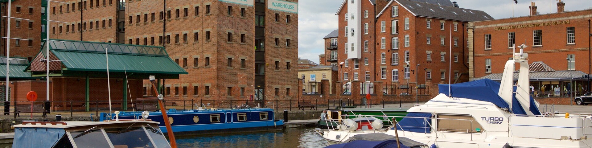 Un puerto deportivo con barcos amarrados, un edificio de ladrillo con el rótulo "VICTORIA WAREHOUSE", y el museo "SOLDIERS OF GLOUCESTERSHIRE MUSEUM" al fondo.