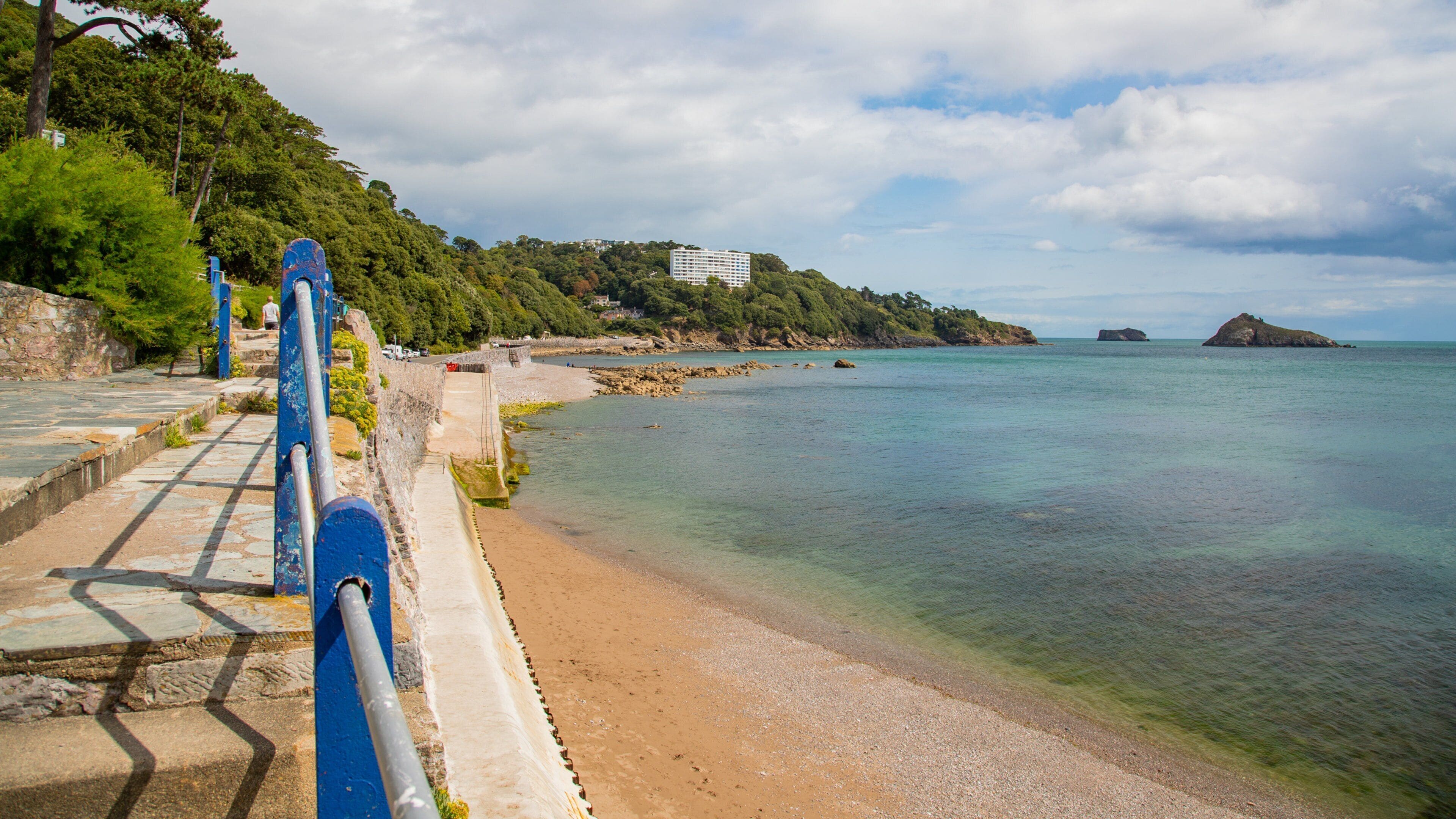 A coastal walkway with a view of the sea, a sandy beach, and a building on a hill.