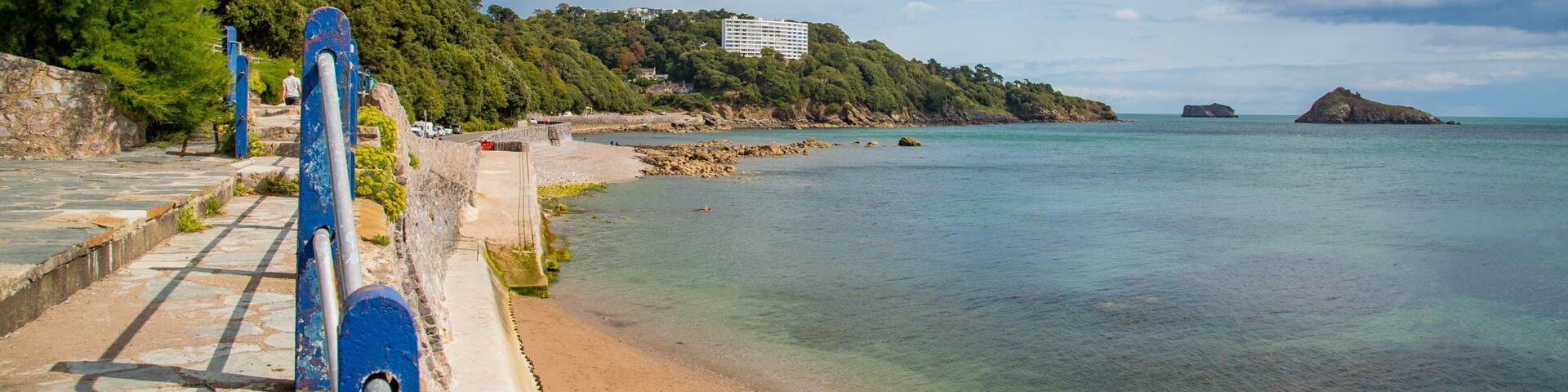 A coastal walkway with a view of the sea, a sandy beach, and a building on a hill.