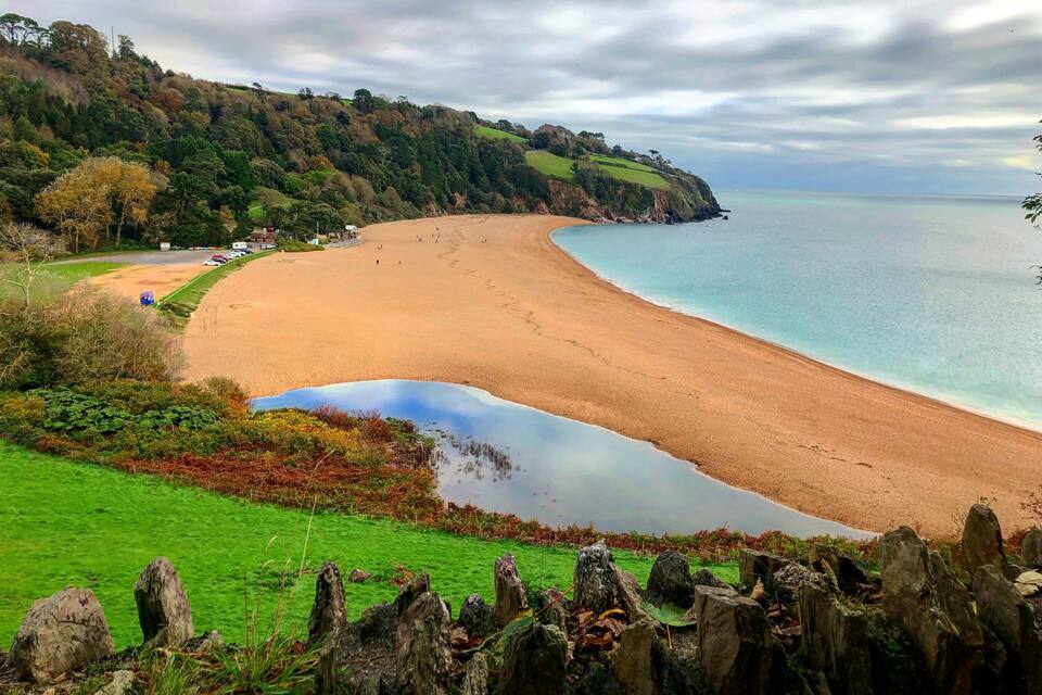Un paisaje costero con playa arenosa, mar tranquilo y una colina verde con árboles.