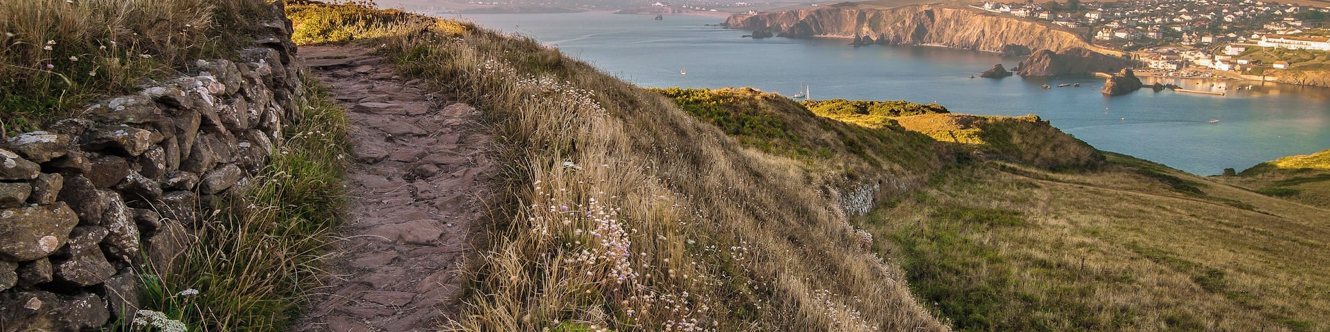 Un paisaje costero con un camino de piedra, colinas cubiertas de hierba y vistas a un pueblo al otro lado del agua.