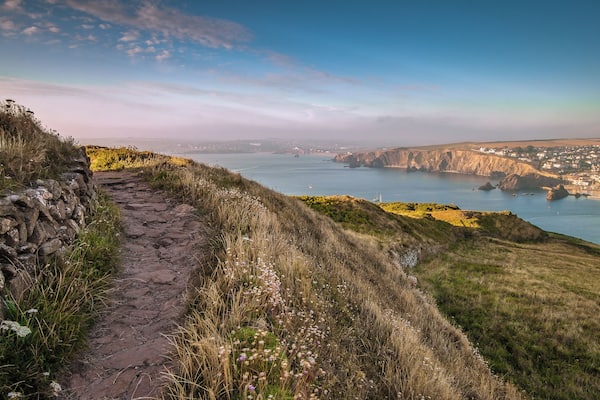 Eine Küstenlandschaft mit einem Steinweg, grünen Hügeln und Blick auf eine Stadt am Wasser.