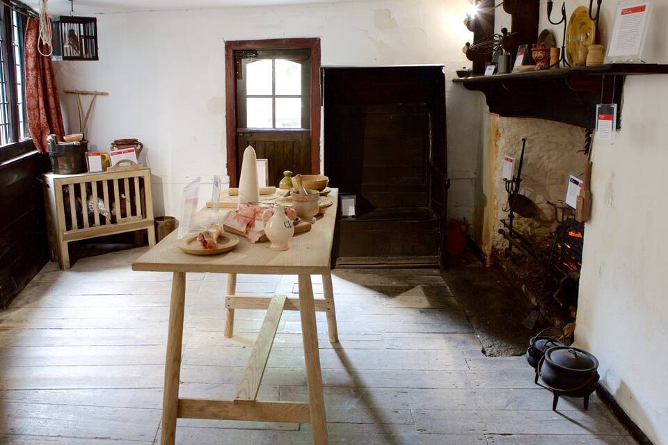 A rustic kitchen with wooden floors, a wooden table with kitchen utensils, a fireplace, and a window with curtains.