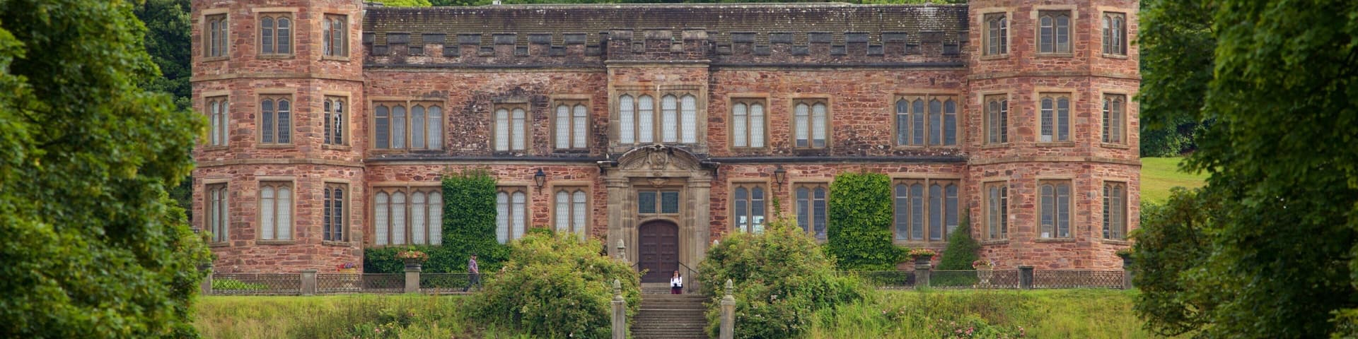 A large, historic brick building with a central entrance and two towers, surrounded by lush greenery.