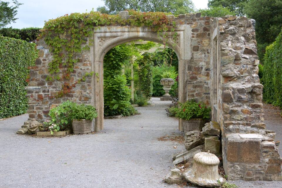 A stone archway with ivy, surrounded by greenery and a gravel path.