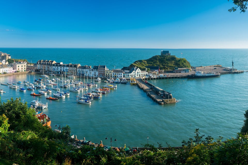 Une ville côtière dotée d’un port de plaisance rempli de bateaux, d’un phare perché sur une colline et d’une structure ressemblant à un château.